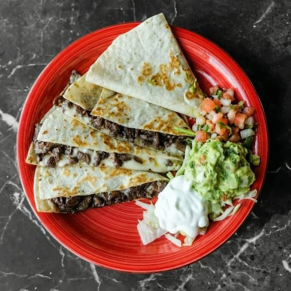 Steak quesadillas with melted cheese, served with guacamole, pico de gallo, and sour cream on a red plate.