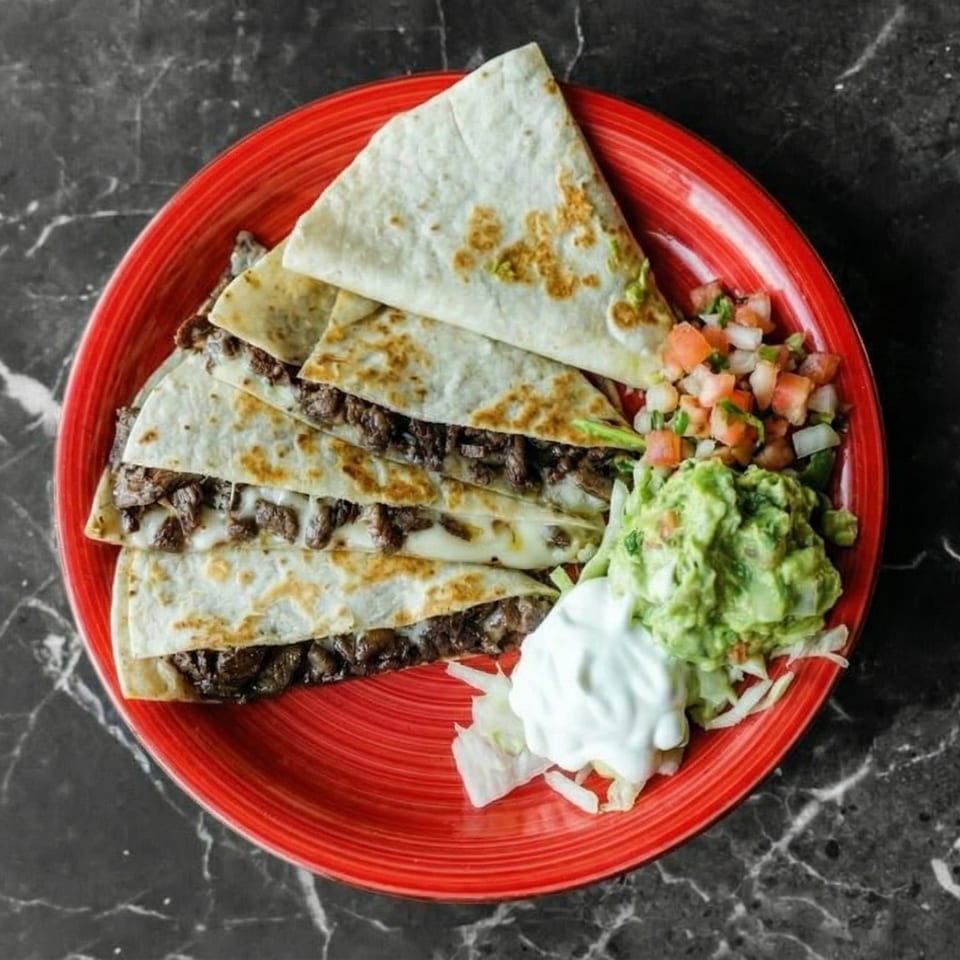 Steak quesadillas with melted cheese, served with guacamole, pico de gallo, and sour cream on a red plate.