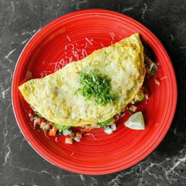A cheesy omelet topped with fresh cilantro, served with pico de gallo and a lime wedge on a red plate.