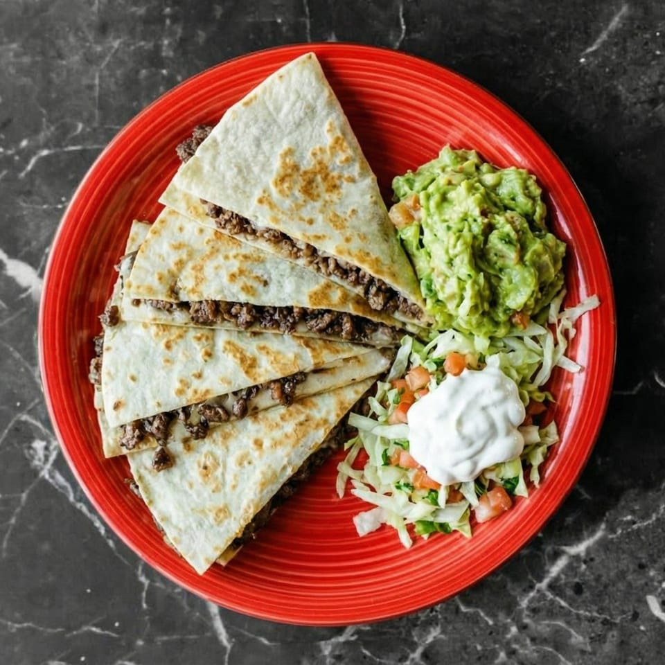 Beef quesadillas with guacamole, shredded lettuce, diced tomatoes, and sour cream on a red plate.